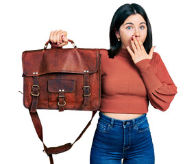 Canvas Print - Young brunette woman with blue eyes holding leather bag covering mouth with hand, shocked and afraid for mistake. surprised expression