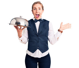 Poster - Young beautiful blonde woman wearing waitress uniform holding tray celebrating victory with happy smile and winner expression with raised hands