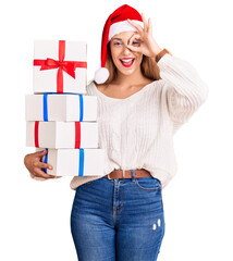 Sticker - Beautiful young woman wearing christmas hat and holding a gift smiling happy doing ok sign with hand on eye looking through fingers