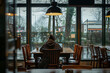 A lone individual sits at a wooden table in the cozy corner of a café, seemingly lost in thought while gazing out of the large glass windows.