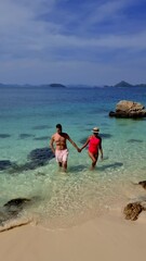 Wall Mural - A diverse couple strolls along the sandy beach, the woman's hand in the man's. The water sparkles under the clear sky, with fluffy clouds drifting above, Ko Kham Thailand