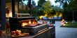 A grill is lit up with a variety of meats and vegetables on it. The grill is surrounded by a patio area with a dining table and chairs. There are several bottles and cups on the table