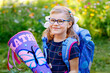 Happy little kid girl with eye glasses with backpack or satchel and big school bag or gift cone traditional in Germany for the first day of school. Healthy adorable child outdoors. Back to school