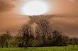 Auf- oder untergehende Sonne mit von Saharastaub orange bis rötlich gefärbtem milchig-trübem Himmel mit Wolken über landwirtschaftlich genutzter Fläche und Bäumen