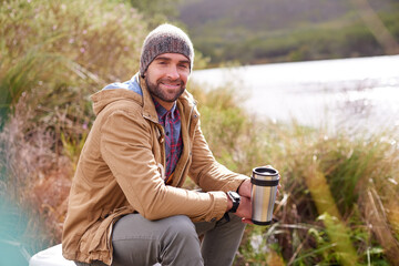 Poster - Camping, portrait and man with coffee by lake in nature for adventure, travel and outdoor vacation. Holiday, smile and male person with hot beverage for relax, peace and winter morning in countryside