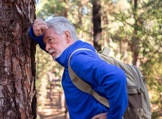 Wall Mural - An active senior man with backpack breathless on a day hike in a mountain forest leans against a tree trunk to regain his strength