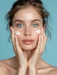 © victoriazarubina - Young woman with a fresh, natural look applying moisturizing cream on her face against a serene blue background. Beauty concept