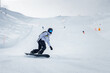 Snowy mountain landscape with winter sports activity. Snowboarder in white jacket and black pants carving turn on slope with snowy spray. Ski resort scene with skiers and lift.