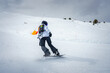 Snowboarder in action on snowy slope, dressed in winter sports gear with bright orange shovel. Winter landscape with snow covered mountains in background.