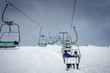 Ski lift taking skiers up snow covered mountain slope with overcast sky. Riders wear helmets, winter gear, chatting or enjoying the ride at a ski resort.