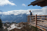 Fototapeta  - Man in casual attire standing on wooden balcony overlooking snow covered mountain village in the European Alps. Majestic snowy peaks in the background.