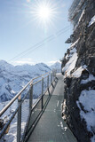 Fototapeta  - Metal walkway on rocky cliff at Murren ski resort, Switzerland. Snow covered mountains in distance, clear blue sky, and safe pathway for exploring Alps.