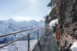 Metal walkway on rocky cliff in Murren ski resort, Switzerland. Mesh structure, railing, snow covered peaks, blue sky. Typical winter setting for skiers.