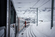 Scenic view of Zermatt ski resort, train journey through snowy landscape. Red or white train rounds bend, snowy mountains, cloud cover, electric lines overhead. Serene winter travel scene.