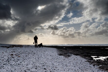 A Man And His Dog Are Walking On A Beach. The Sky Is Cloudy And The Beach Is Covered In Snow