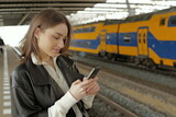 Fototapeta Na ścianę - Portrait of a young woman using mobile phone waiting for a train at a station 
