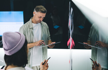 Fototapete - Caucasian male in bluetooth earbuds using digital tablet while collaborate with female colleague during brainstorming in office interior, professional employees working together on startup project