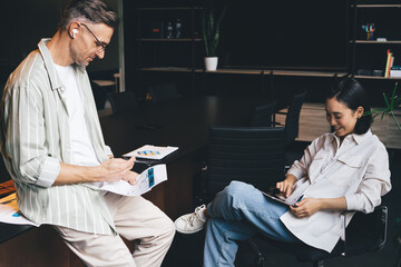 Fototapete - Asian female searching web content via application on touch pad during collaborative working process with male colleague in bluetooth earbuds, professional partners discussing business marketing