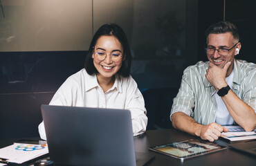 Fototapete - Happy male and female IT professionals reading publication about UI UX graphic design for web product and smiling at table desktop with modern laptop computer, cheerful colleagues collaborating