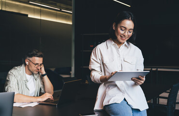Fototapete - Happy Asian employee in classic eyewear for vision correction using digital tablet for browsing information on website via application, successful graphic designer in spectacles making online booking