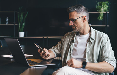 Fototapete - Side view of Caucasian male executive manager siting at desktop with modern laptop computer and using cellphone gadget for browsing website, skilled businessman in spectacles checking mobile email