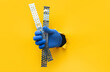 A rsght man's hand in a blue knitted glove holds a U-shaped fastening profile for drywall. Torn hole in yellow paper. The concept of a worker, a labor migrant, a master of his craft. Copy space.