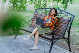 Fototapeta Mapy - Smiling asian woman talking on speakerphone on smartphone while sitting with laptop on city park bench. Lifestyle freelancer, woman negotiating in park on smartphone outdoors in park.