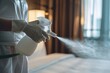 A housekeeper in a white uniform sprays disinfectant on a bed in a well-lit hotel room, ensuring cleanliness. Housekeeping Staff Disinfecting Hotel Room
