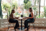 Fototapeta Na drzwi - Two women enjoy collaborative work in tropical café. woman with braids smiles as she listens, while her companion gestures during conversation. laptop and note-taking indicate productive atmosphere
