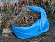 An open  blue garbage bag full of dry branches, grass and plants standing outdoors.