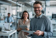 © Kien - Portrait of a happy man with a digital tablet standing in front of his female coworker working at the office, laughing and smiling while looking at the camera