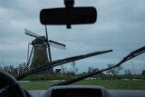 Fototapeta Miasto - A typically miserable rainy day in the Netherlands view from inside a car. windmill and grey stormy clouds with rear view mirror, dashboard and windscreen wipers on Dutch bad weather day in Holland