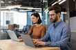 © Liubomir - Focused man and woman using laptops at a wooden table in a bright, contemporary workspace, embodying professionalism and teamwork.