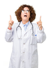 Canvas Print - Middle ager senior doctor woman over isolated background amazed and surprised looking up and pointing with fingers and raised arms.