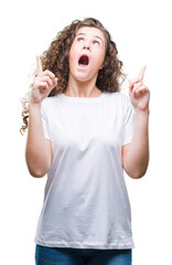 Wall Mural - Beautiful brunette curly hair young girl wearing casual t-shirt over isolated background amazed and surprised looking up and pointing with fingers and raised arms.