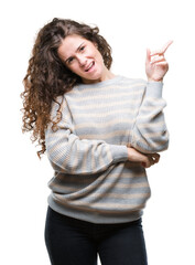 Poster - Beautiful brunette curly hair young girl wearing winter sweater over isolated background with a big smile on face, pointing with hand and finger to the side looking at the camera.