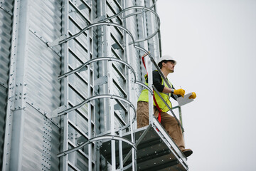 A male industrial worker stands on a grain silo with documents in his hands and makes a visual inspection.