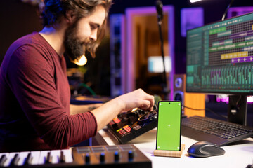 Poster - Music producer uses controls and knobs on mixing console in home studio, having a smartphone with greenscreen display. Artist learning to edit tunes and add sound effects with daw software.