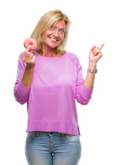 Poster - Middle age blonde woman eating pink donut over isolated background very happy pointing with hand and finger to the side