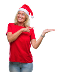 Canvas Print - Middle age blonde woman wearing christmas hat over isolated background amazed and smiling to the camera while presenting with hand and pointing with finger.