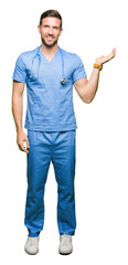 Wall Mural - Handsome doctor man wearing medical uniform over isolated background smiling cheerful presenting and pointing with palm of hand looking at the camera.