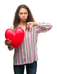 Poster - Young hispanic woman in love holding red heart with angry face, negative sign showing dislike with thumbs down, rejection concept