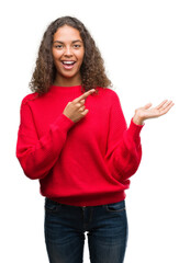 Canvas Print - Young hispanic woman wearing red sweater amazed and smiling to the camera while presenting with hand and pointing with finger.