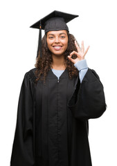 Wall Mural - Young hispanic woman wearing graduation uniform doing ok sign with fingers, excellent symbol