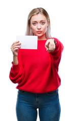 Wall Mural - Young blonde woman holding blank paper card over isolated background pointing with finger to the camera and to you, hand sign, positive and confident gesture from the front