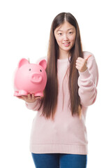 Poster - Young Chinese woman over isolated background holding piggy bank happy with big smile doing ok sign, thumb up with fingers, excellent sign