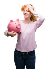 Poster - Young redhead woman holding piggy bank stressed with hand on head, shocked with shame and surprise face, angry and frustrated. Fear and upset for mistake.