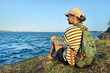 rear view Young woman wearing cap with backpack sitting on rocks and looking at view of ocean
