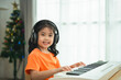 Asian young girl is smiling sitting at a piano, wearing headphones. She is wearing an orange shirt. The room is filled with books and a keyboard