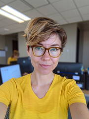 Wall Mural - A selfie of an average looking woman with short hair and glasses, wearing yellow top in her office. It feels like she was taking it while working, a computer screen out of focus in background